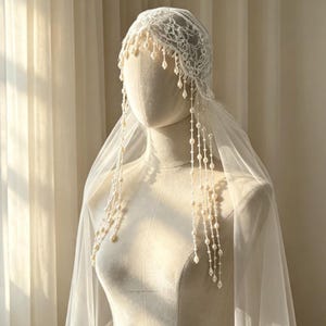 May include: A white lace cap veil featuring intricate floral patterns and delicate hanging strands of white pearls. The headpiece includes a sheer, flowing tulle veil that drapes over the shoulders, creating a layered and elegant appearance.