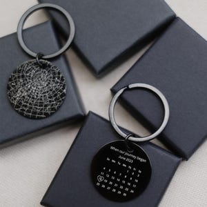 May include: Two black keychains with round pendants. One pendant features a web design, the other displays a calendar for June 2023 with the text "When our journey began". The keychains are on black boxes.