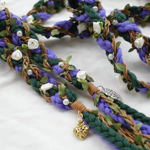 Könnte beinhalten: Geflochtenes Haaraccessoire mit lila, grünen und braunen Strähnen, verziert mit weißen Rosen, Kunstperlen und grünen Blättern. Ein goldener Herzanhänger und ein silberner Anhänger fügen Details hinzu.