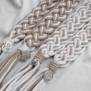 May include: Close-up of four macrame braids in neutral tones of white, beige, and brown. Each braid features a different silver charm, including an infinity symbol, a tree of life, and a heart. The braids are made of thick, woven cotton rope.