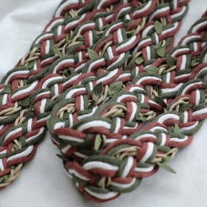 Puede incluir: Primer plano de un artículo decorativo trenzado con un patrón repetitivo de hebras rojas, blancas, verdes y beige. Pequeños acentos de hojas verdes se intercalan en la trenza. El artículo es probablemente hecho a mano y descansa sobre una superficie blanca.