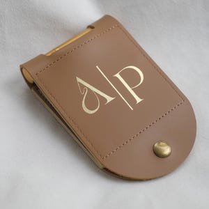 Op de afbeelding: Een bruinleren portemonnee met goudkleurige hardware en een drukknoopsluiting. De portemonnee heeft de goudkleurige initialen "A P" op de voorkant.