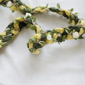 May include: A braided floral crown with yellow and green yarn, adorned with small white flowers and green leaves. The crown is designed with a natural, rustic aesthetic, perfect for weddings or special events.