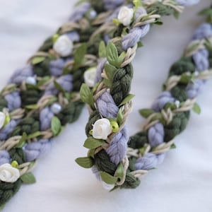 Könnte beinhalten: Geflochtenes Blumen-Haarband mit grüner, lavendelfarbener und beiger Wolle. Das Design ist mit kleinen weißen Rosen, grünen Blättern und winzigen weißen Perlen verziert, was eine zarte und natürliche Ästhetik erzeugt.