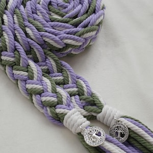 May include: Braided macrame rope in shades of lavender, green, and white, with silver tree of life charms. The rope is coiled and braided, creating a textured pattern. The charms are attached to the ends of the braided rope.