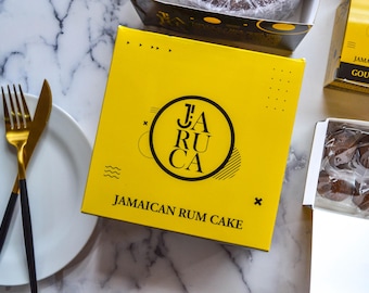 以下が含まれることがあります： 「JA RU CA JAMAICAN RUM CAKE」と黒文字で書かれた黄色の箱。箱は白い大理石の表面に置かれている。表面には白い皿と2本の金色のフォークがある。