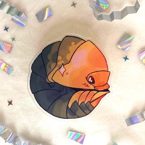 Pode incluir: Um autocolante colorido com um inseto de desenho animado enrolado, com cabeça laranja, corpo castanho e barriga azul escura. O autocolante tem um efeito brilhante. Confetes holográficos e confetes em forma de estrela espalhados.