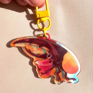 Puede incluir: Un llavero acrílico colorido con un escarabajo de dibujos animados sosteniendo un pequeño cubo rojo. El llavero tiene un cierre y un anillo amarillos. El escarabajo tiene un cuerpo marrón, un ala naranja y un cuerno grande. El fondo tiene tonos naranja y azul.