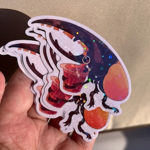 Op de afbeelding: Een stapel holografische stickers met een cartoonkever met een rode beker. De kever heeft een donkerblauwe kop met oranje accenten en een sterrenontwerp. De stickers worden in een hand gehouden.