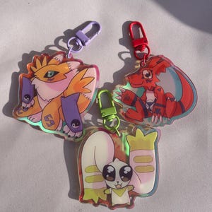 Peut inclure: Trois porte-clés colorés représentant des personnages de dessins animés de la série d'anime Digimon. Les porte-clés sont en acrylique transparent avec une finition holographique. Les personnages sont une créature ressemblant à un renard jaune et orange, une créature ressemblant à un dinosaure rouge et orange, et une créature ressemblant à un lapin blanc et jaune.