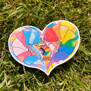 Puede incluir: Una pegatina en forma de corazón con dos figuras de dibujos animados que se toman de la mano. Las figuras están en los colores de la bandera arcoíris y el corazón está lleno de un arcoíris y rayas negras y marrones. El texto "Love is Love" está escrito en el centro del corazón.