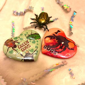 May include: A collection of bug-themed items. A metallic gold beetle figurine is positioned above two heart-shaped badges. One badge is green with the text "GREEN GUYS" and bug illustrations. The other is red with a bug illustration and the text "I BUGS".