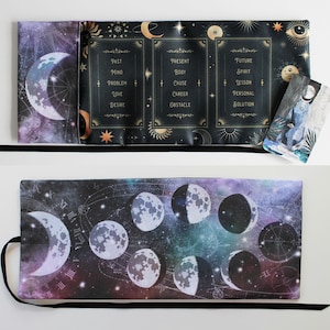 May include: A black tarot card mat with a celestial design featuring moons, stars, and text. The mat is open, revealing three sections with words like "Past," "Present," and "Future." A tarot card with a blue figure is on the mat.