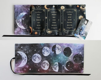 May include: A black tarot card mat with a celestial design featuring moons, stars, and text. The mat is open, revealing three sections with words like "Past," "Present," and "Future." A tarot card with a blue figure is on the mat.
