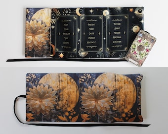 May include: A celestial-themed tarot card holder and mat. The mat features a large, stylized flower and a moon design in shades of gold and brown against a dark blue background. The holder has text that reads "Past, Present, Future". A single tarot card is included.
