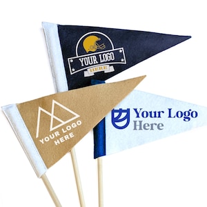 Puede incluir: Tres banderas triangulares en palos de madera. Una es azul marino con un gráfico de casco de fútbol y el texto "YOUR LOGO HERE". Otra es de color canela con un logotipo de montaña y el texto "YOUR LOGO HERE". La tercera es blanca con un logotipo azul y el texto "Your Logo Here".