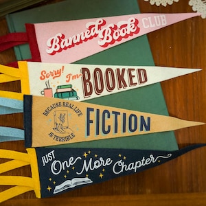 May include: Four felt pennants with book-themed phrases. The pennants are pink, cream, tan, and navy blue. Text includes "Banned Book Club", "Sorry! I'm Booked", "Because real life is terrible, fiction", and "Just One More Chapter".
