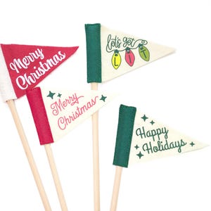 Könnte beinhalten: Eine Sammlung festlicher Wimpel mit Holzstäben. Die Flaggen zeigen Weihnachtsgrüße wie "Merry Christmas" und "Happy Holidays" in weißer Schrift auf rotem und cremefarbenem Filz. Eine Flagge sagt "let's get lit" mit Lichterketten.