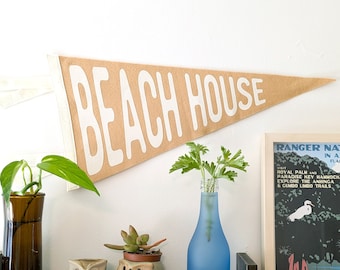 May include: A tan felt pennant with the text "BEACH HOUSE" in white lettering. The pennant is hanging on a white wall. There are three potted plants, a wooden shelf, and a framed poster with text about a nature preserve.