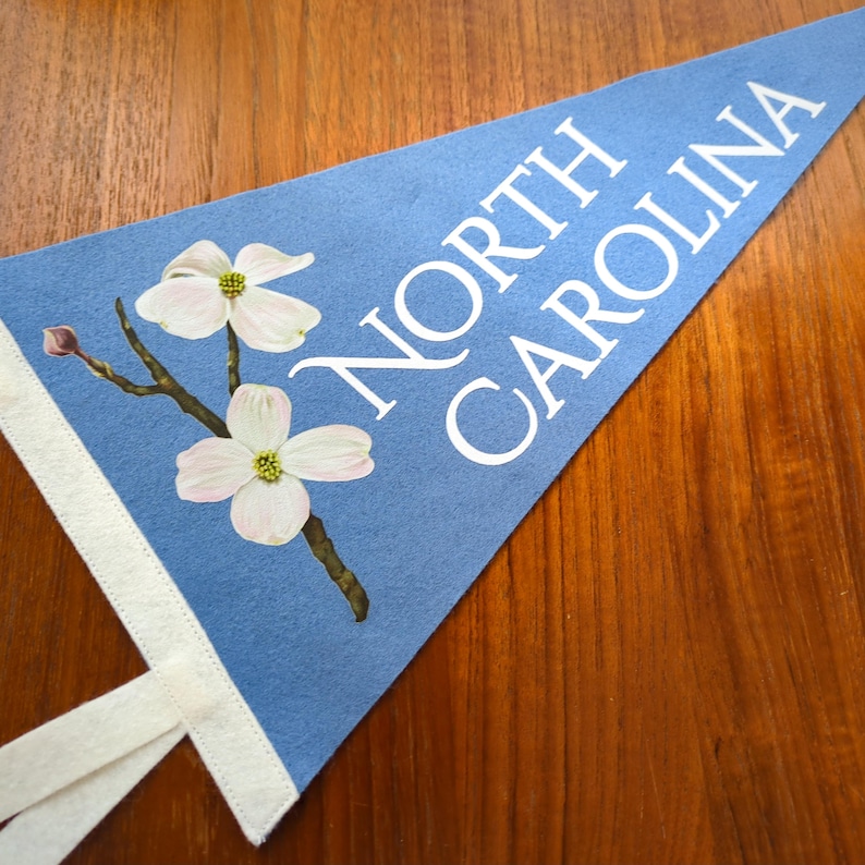 May include: A light blue felt pennant with the words "NORTH CAROLINA" in white. The pennant features a floral design with white and pink dogwood blossoms. A white ribbon is attached to the pennant's edge.