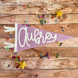 Peut inclure: Une banni&egrave;re en feutre violet avec le nom Aubrey &eacute;crit en lettres cursives blanches. La banni&egrave;re est d&eacute;cor&eacute;e de franges blanches et est entour&eacute;e de pompons color&eacute;s sur un fond en bois.
