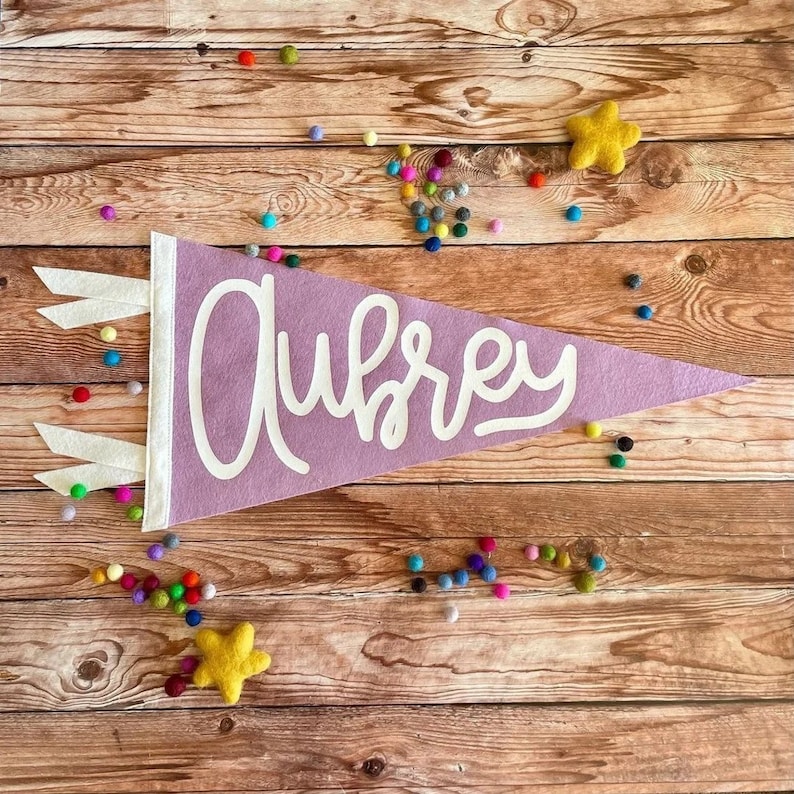 Peut inclure: Une banni&egrave;re en feutre violet avec le nom Aubrey &eacute;crit en lettres cursives blanches. La banni&egrave;re est d&eacute;cor&eacute;e de franges blanches et est entour&eacute;e de pompons color&eacute;s sur un fond en bois.