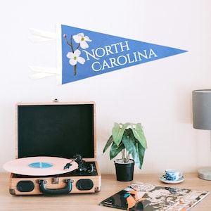 May include: A blue pennant with the text "NORTH CAROLINA" and a floral design. A vintage record player with a pink record is on a wooden table, along with a small potted plant, a teacup, and a lamp.