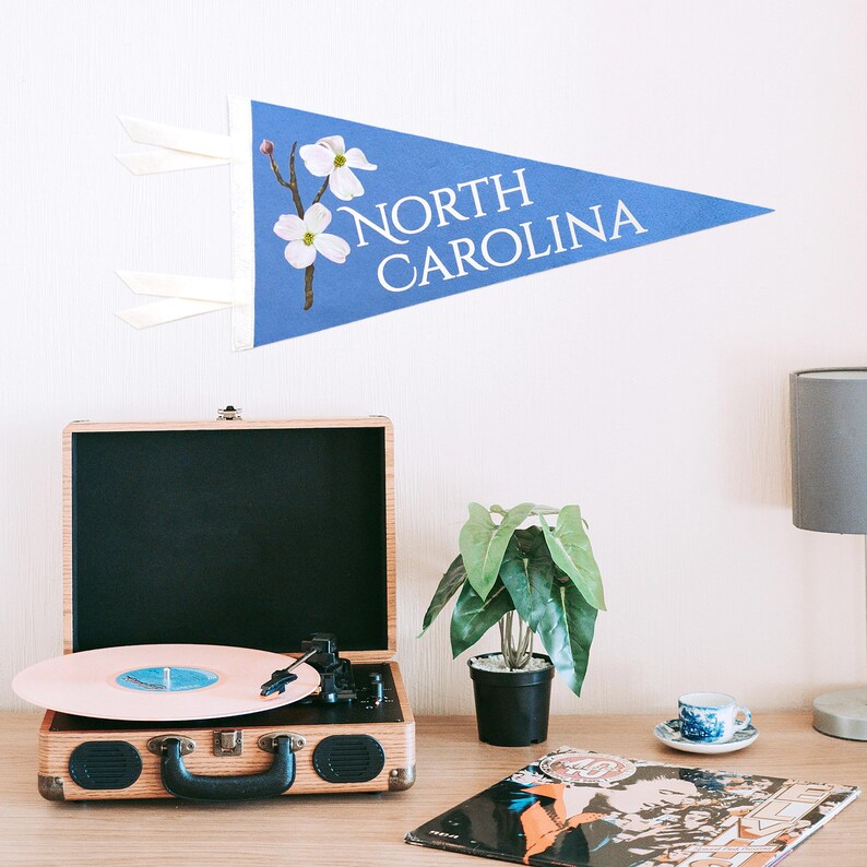 May include: A blue pennant with the text "NORTH CAROLINA" and a floral design. A vintage record player with a pink record is on a wooden table, along with a small potted plant, a teacup, and a lamp.