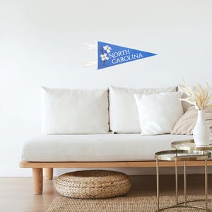 May include: A blue pennant with the words "NORTH CAROLINA" and a floral design. The pennant is hanging on a white wall above a white couch with pillows. The room also features a woven ottoman and gold side tables.