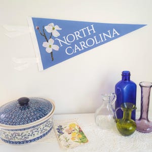 May include: A light blue felt pennant with the words "NORTH CAROLINA" in white. The pennant features a floral design with white flowers and a brown stem. White ribbons are attached to the pennant.
