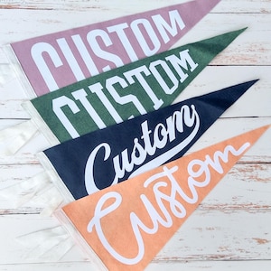 May include: Four triangular felt pennants in dusty pink, forest green, navy blue, and peach. Each pennant features the word CUSTOM printed in white, with the top two in a block font and the bottom two in different styles of white cursive script.
