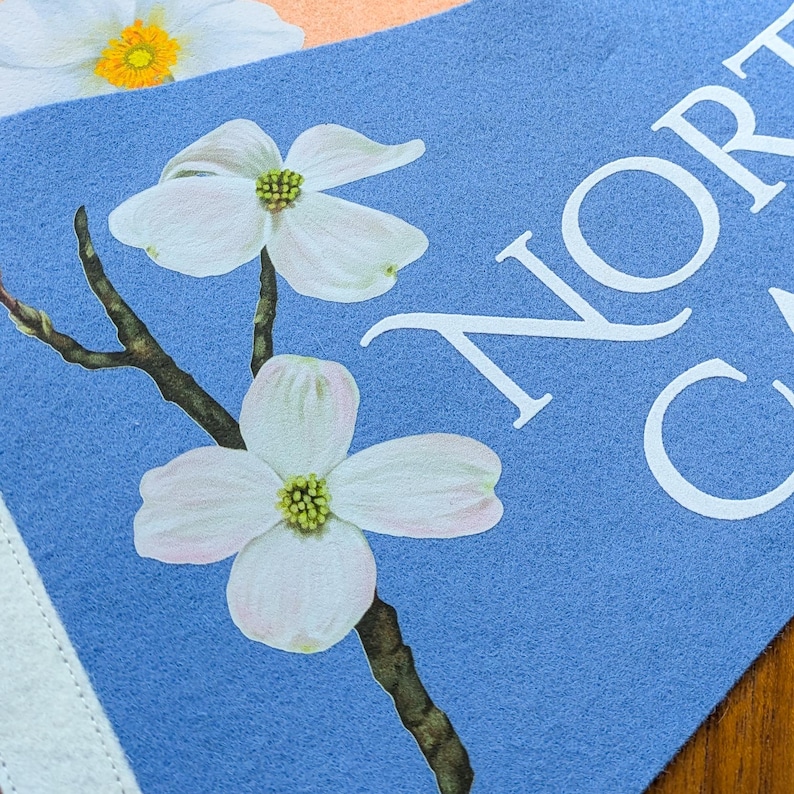 May include: A blue felt pennant with the text "NORTH CAROLINA" in white. The pennant features a detailed illustration of white dogwood flowers with yellow centers and a brown branch. The background is a light blue color.