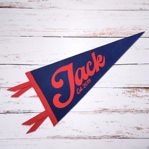 Peut inclure: Un fanion bleu en feutre avec une bordure rouge et le nom "Jack" en lettres rouges cursives. Le fanion comprend &eacute;galement le texte "Est. 2019" en rouge.