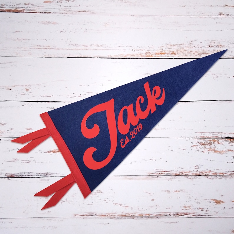 Peut inclure: Un fanion bleu en feutre avec une bordure rouge et le nom "Jack" en lettres rouges cursives. Le fanion comprend &eacute;galement le texte "Est. 2019" en rouge.