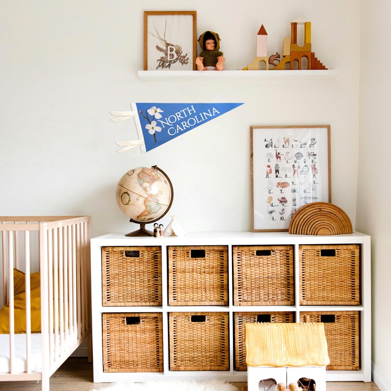May include: A nursery room with a white crib, a white storage unit with woven baskets, and a blue pennant that says "NORTH CAROLINA". A globe and framed art pieces are also visible.