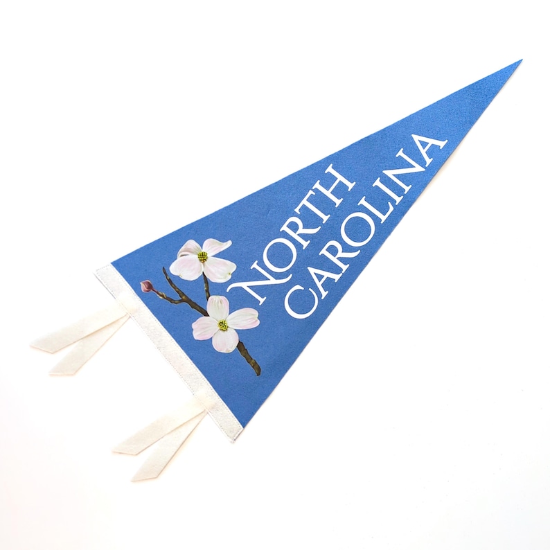 May include: A blue pennant with the words "NORTH CAROLINA" in white. The pennant features a floral design with white flowers and a brown branch. White ribbons are attached to the pennant.