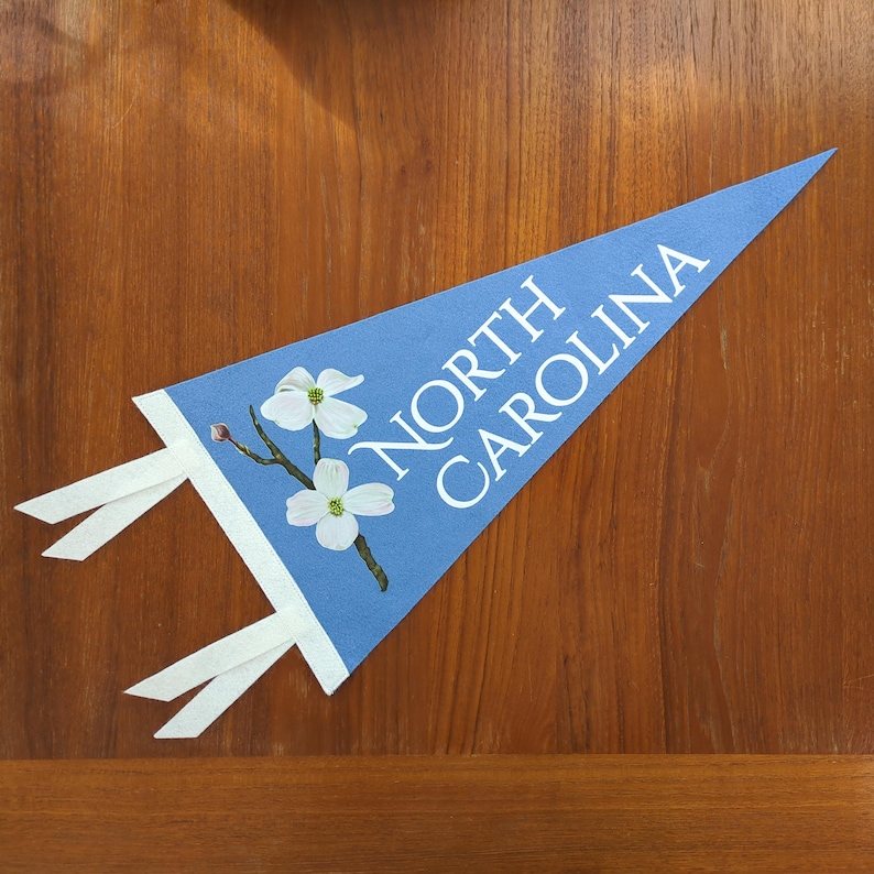 May include: A light blue pennant with the words "NORTH CAROLINA" in white. The pennant features a white border and a floral illustration of white dogwood blossoms. White ribbons extend from the pennant's base.
