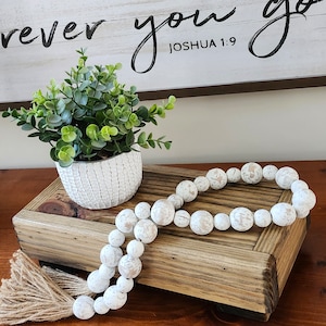 May include: A wooden tiered stand with a white beaded garland and a potted green plant. The stand has a distressed wood finish and the garland is made of white beads with a speckled finish. The plant is in a white ceramic pot with a textured surface. The stand is on a wooden surface and the text "Wherever you go Joshua 1:9" is visible in the background.