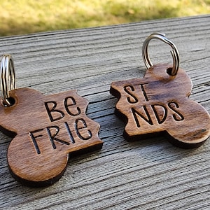 May include: Two wooden keychains with a brown stain. The keychains are shaped like puzzle pieces and say "Be Friends" on one and "Best" on the other.