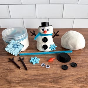 May include: A build your own snowman kit featuring a clear plastic container of white modeling clay, a finished snowman figure, a ball of white clay, two black buttons, two googly eyes, a black hat, a carrot-shaped nose, and blue snowflake.