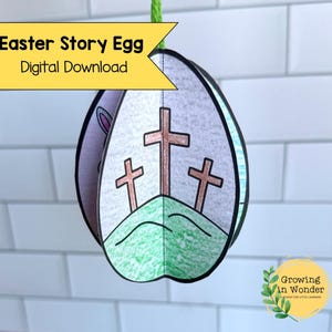 Può includere: Un uovo di Pasqua di carta, download digitale, con tre croci su una collina verde. L'uovo è sospeso da uno spago verde. Il testo "Easter Story Egg Digital Download" è visualizzato su uno striscione giallo.