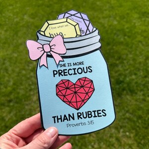 May include: A light blue mason jar-shaped card with the text "SHE IS MORE PRECIOUS THAN RUBIES" and "Proverbs 3:15". It features a pink bow and a red heart. Inside the jar are three gem-shaped cards with text.