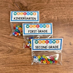 Könnte beinhalten: Drei transparente Beutel mit bunten Perlen, jeweils mit "WELCOME" und einer Klassenstufe beschriftet: Kindergarten, 1. Klasse und 2. Klasse. Die Perlen sind in verschiedenen Farben, darunter Blau, Orange, Grün und Weiß.