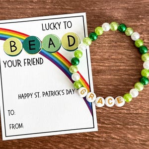 May include: A beaded bracelet with green, white, and gold beads next to a card. The card has a rainbow and text that reads "Lucky to Bead Your Friend" and "Happy St. Patrick's Day". The bracelet has the name "Grace".