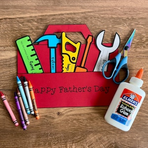 May include: A red paper toolbox with cut-out tools and the text "Happy Father's Day." Includes crayons and a bottle of school glue. The toolbox contains a ruler, hammer, saw, wrench, and scissors.