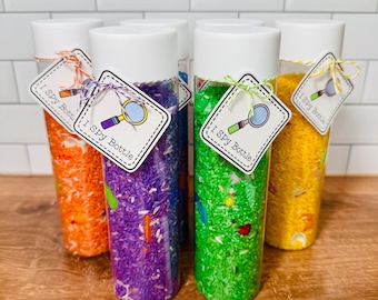 May include: Six clear plastic sensory bottles filled with colorful rice and small objects. Each bottle has a white lid and a tag that reads "I Spy Bottle" with a magnifying glass illustration. The rice is in shades of orange, purple, green, blue, and yellow.