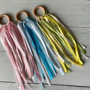 May include: Three wooden rings with colorful ribbons attached. The rings are in shades of brown and the ribbons are pink, blue, and yellow.