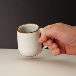 May include: A white ceramic mug with a brown rim and a handle. The mug is being held by a hand.