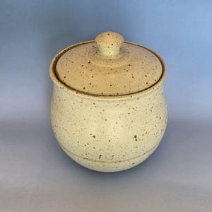 Puede incluir: Un tarro de cer&aacute;mica moteado con tapa. El tarro es de color blanquecino con motas marrones y tiene forma redondeada. La tapa tiene un peque&ntilde;o pomo en la parte superior. El tarro se utiliza probablemente para guardar cosas o para decoraci&oacute;n.