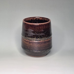 May include: A handmade ceramic tumbler with a rich, dark brown glaze. The cup features a cylindrical shape with a slightly flared top and a band of lighter brown around the middle. The glaze has a speckled appearance, adding texture and visual interest.